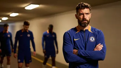 A 35-year-old Chelsea FC assistant coach, a ruggedly built man with a thick beard and a stern expression, stands in the...