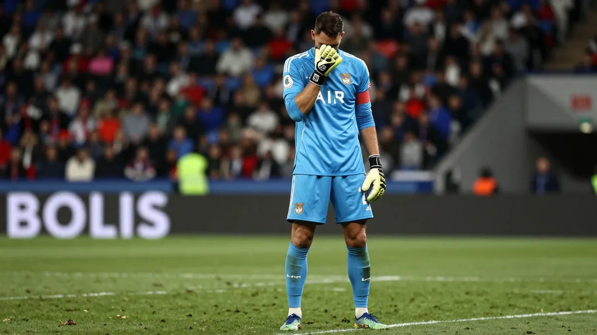 A goalkeeper in a blue kit standing on a grass pitch looking de dejected, blurred stadium background