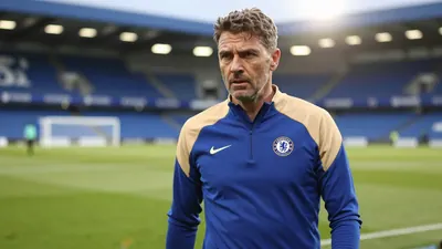 A 35-year-old Chelsea FC assistant coach with a rugged build and weathered face, dressed in a plain blue and amber...