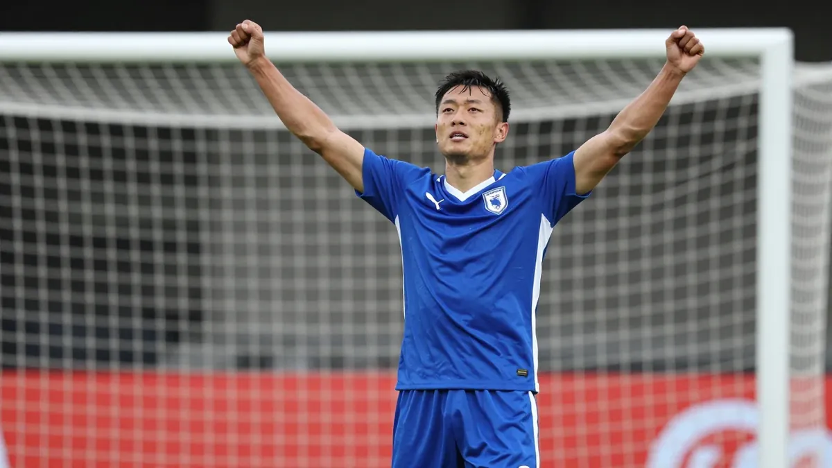 A 30-year-old male football player, wearing a blue jersey, celebrating a goal with his arms raised, photographed with a...